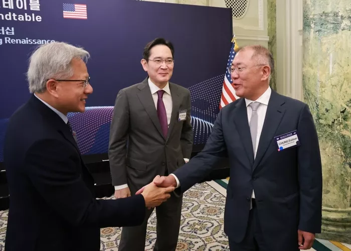 Three men in suits shake hands in a conference room labeled “Roundtable,” with American and South Korean flags in the background.