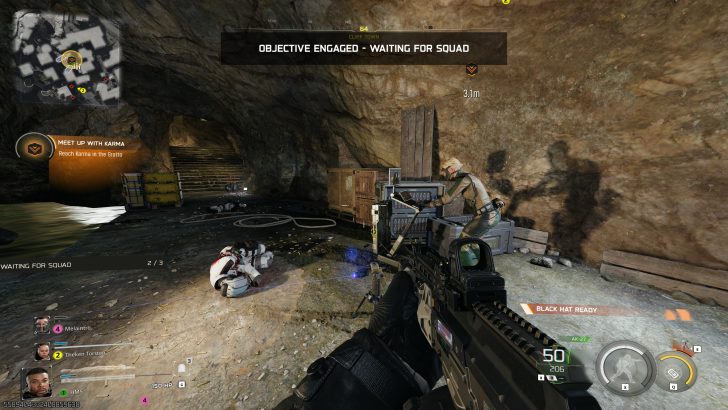 A first-person shooter game screen shows a character aiming a rifle inside a cave with the objectives 'Meet Up with Karma' and 'Objective Engaged - Waiting for Squad' displayed.