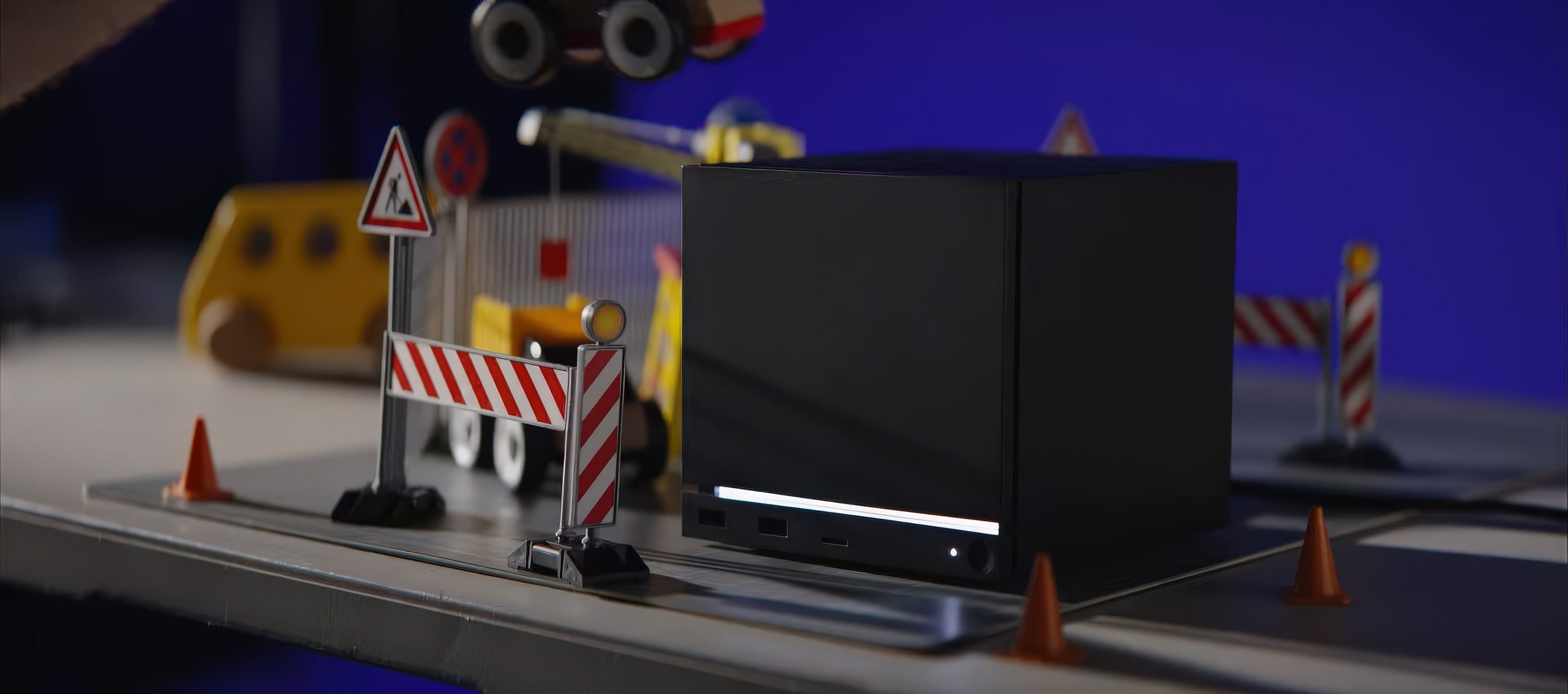 A black cube-like device sits on a table surrounded by toy traffic cones, construction barriers, and miniature vehicles against a blue background.