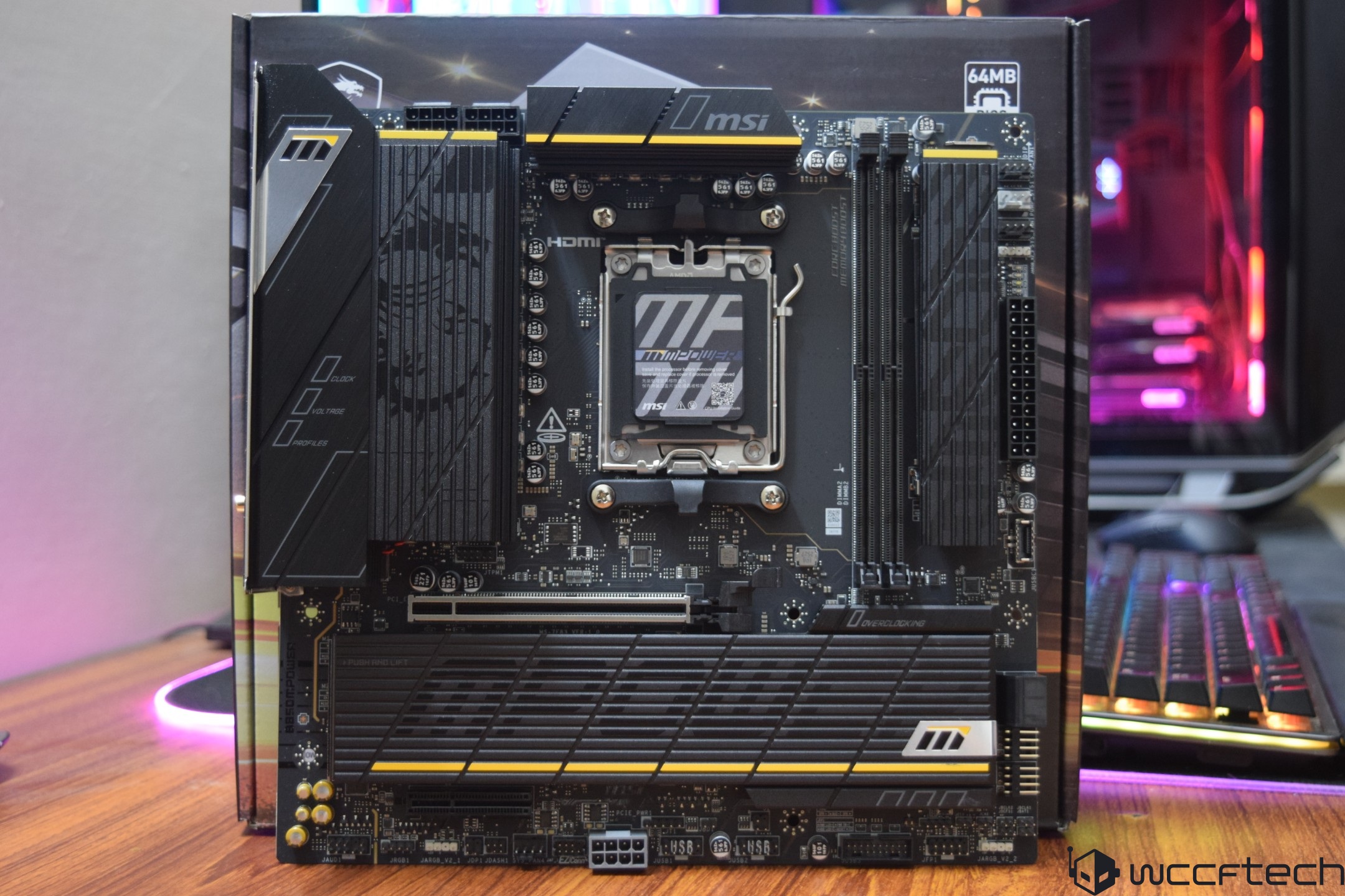 MSI motherboard with DDR5, 64MB, and Overclocking labels displayed on a wooden table in front of a colorful keyboard.