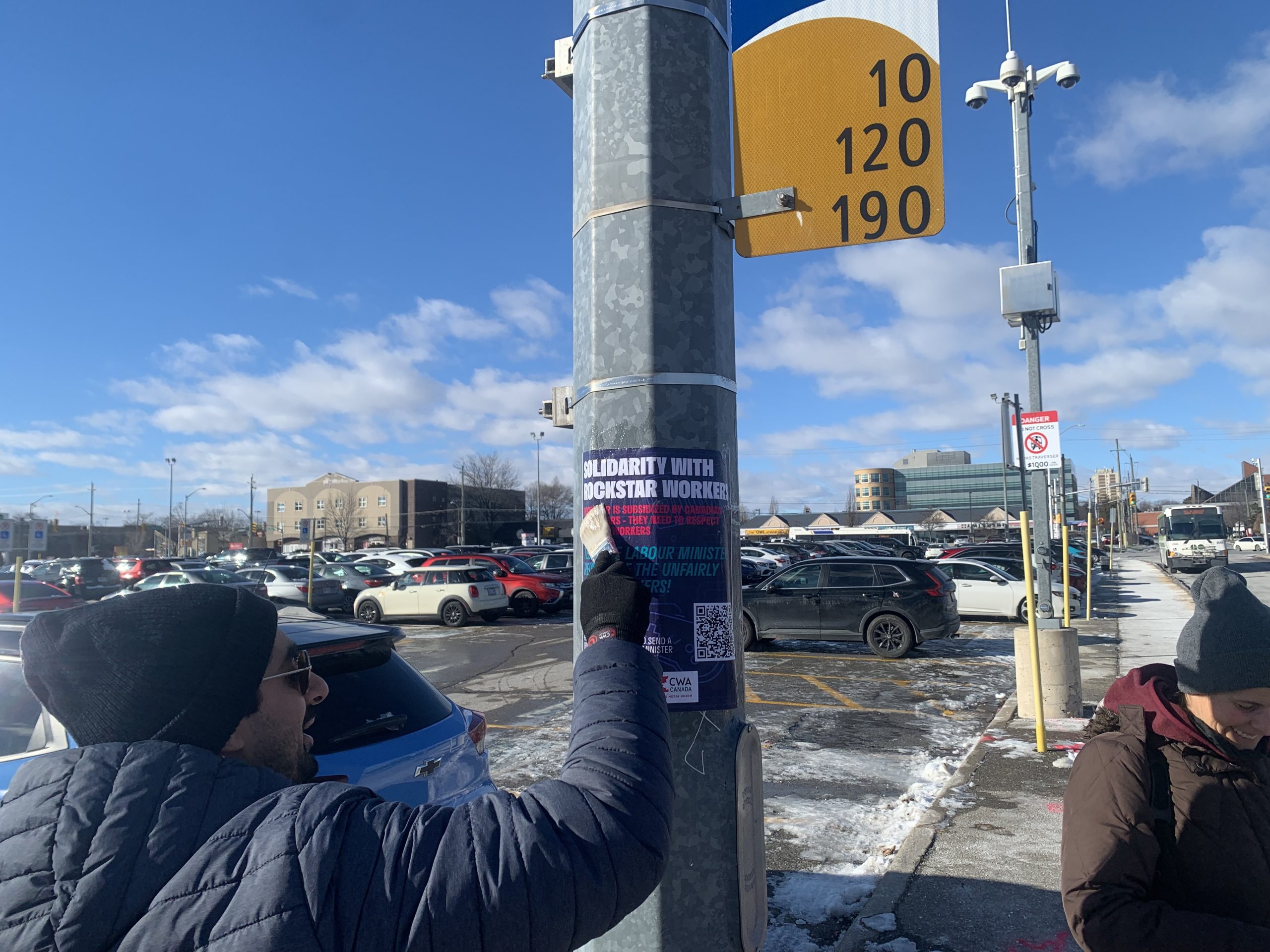 A person wearing a winter jacket and beanie is removing a poster titled 'Solidarity with Rockstar Workers' from a snow-dusted parking lot signpost.