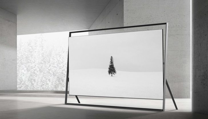 A large, thin-framed television displaying an image of a solitary tree on a snowy landscape stands in a modern interior with concrete walls and a snowy window view.